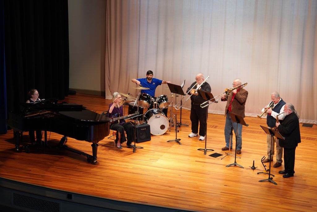 group of musicians at a piano, drum set, keyboard, and with saxophone, trombones and trumpets playing in an auditorium