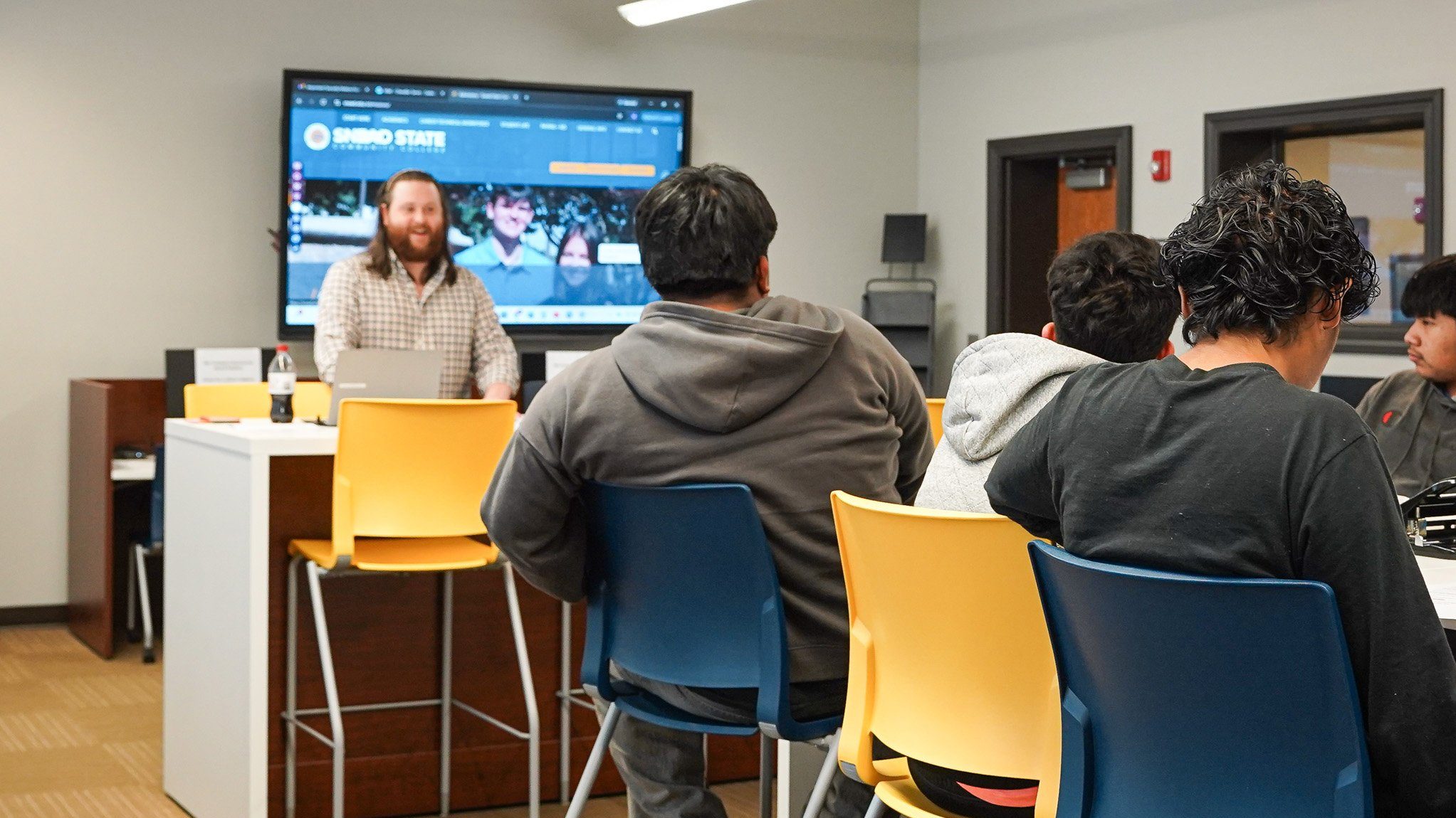 high school students sitting at a table with a college representative standing in front with a presentation on a big video screen behind him