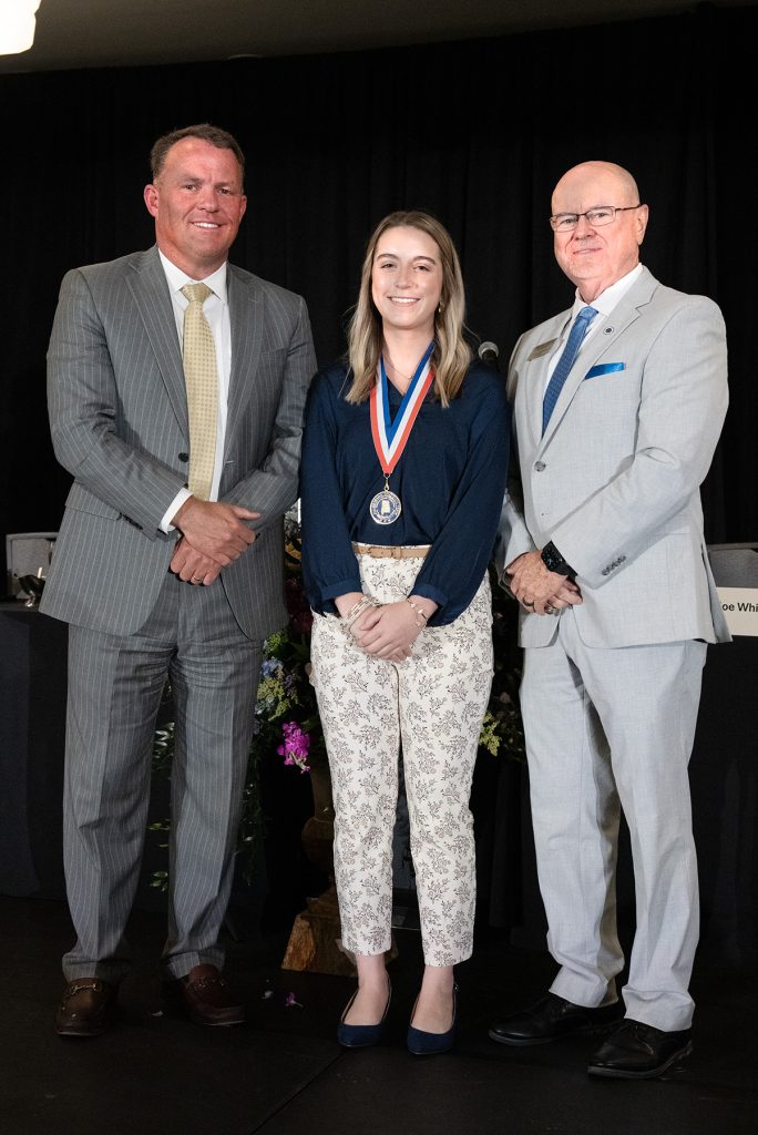 two men wearing suits stand on either side of a female student wearing an award medallion